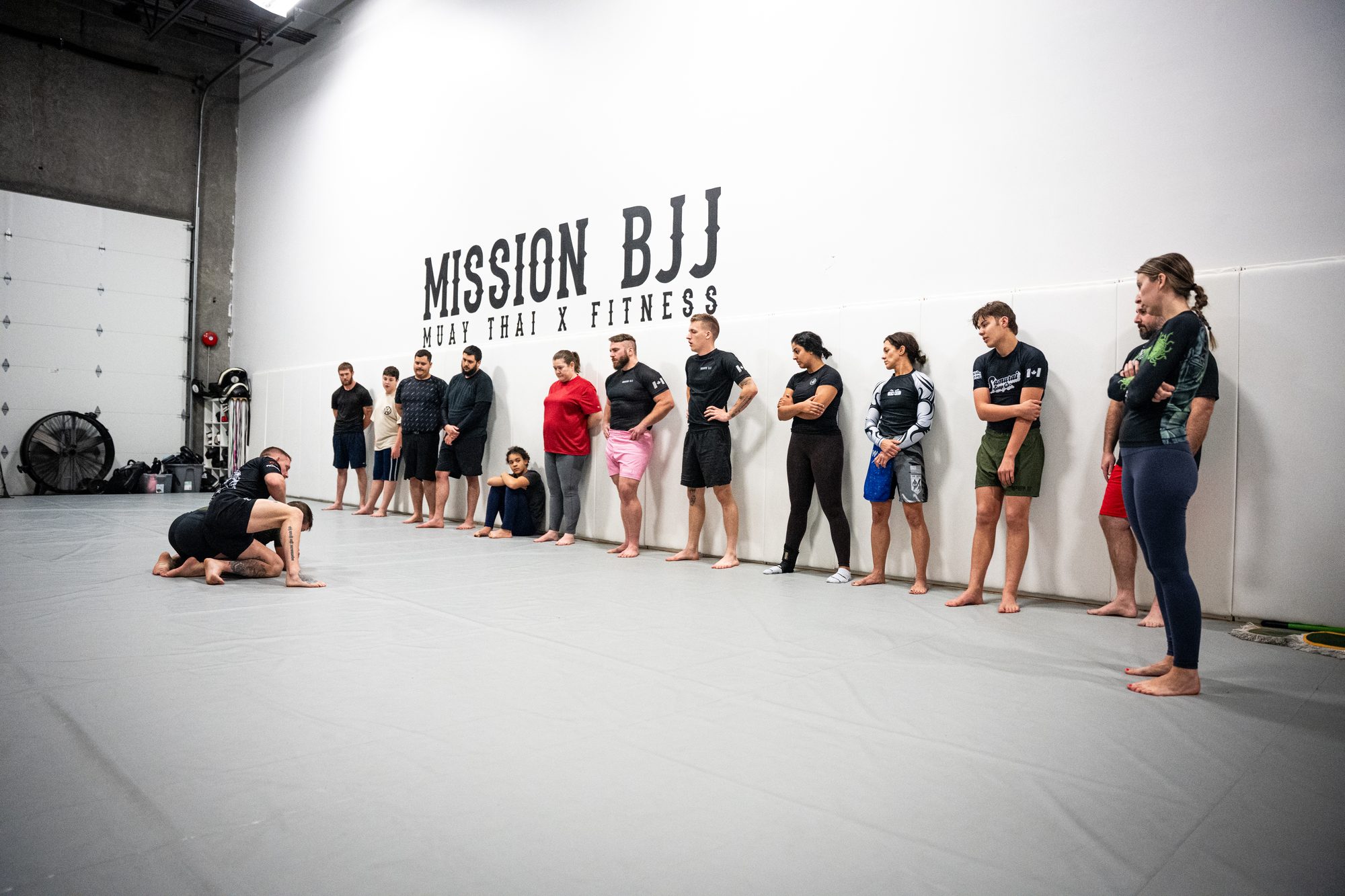 Class lined up beneath the painted Mission BJJ wall logo as a coach demonstrates a movement on the mats in Mission, BC